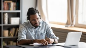 man writing notes from his laptop with headset on