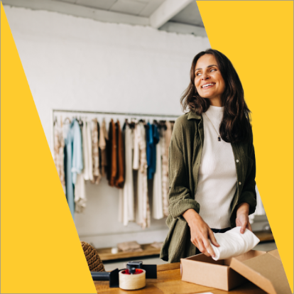 A woman smiling while folding a white garment into a cardboard box in a clothing store. A rack of clothes is visible in the background, symbolizing affordable small business shipping solutions that cater to diverse needs.