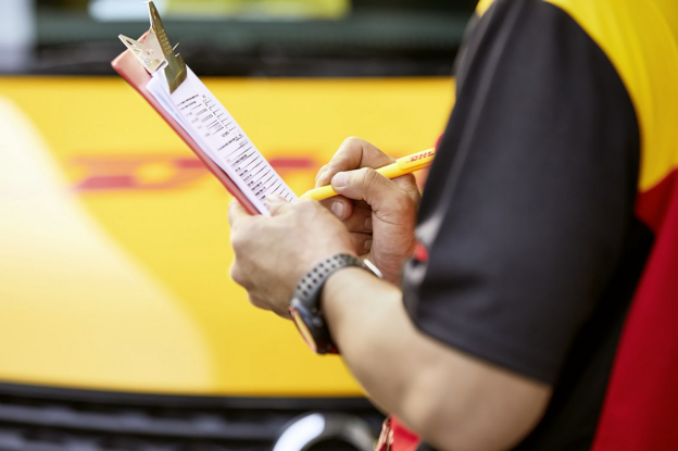 A person wearing a black and red uniform writes on a clipboard with a yellow pen in front of a yellow vehicle.