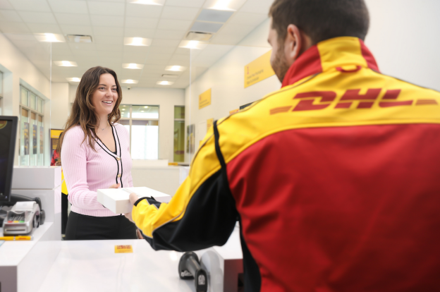 A woman at a counter hands a package to a person wearing a DHL jacket in a brightly lit office setting, highlighting the convenience of shipping for small businesses looking to expand internationally.