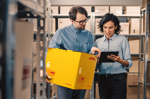 Two individuals in a warehouse setting; one holds a yellow DHL box while the other points at a tablet screen, likely discussing affordable small business shipping solutions.