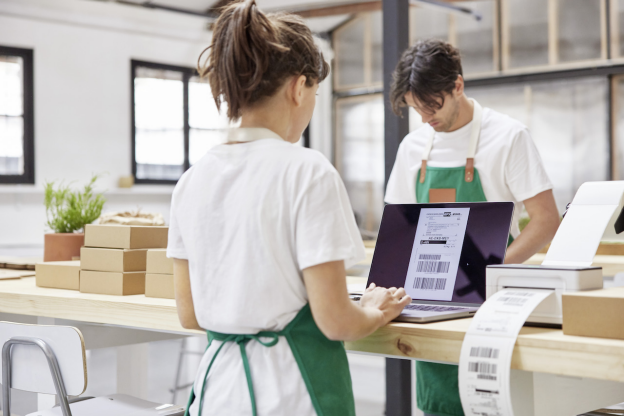 Two people wearing green aprons work in a warehouse. The person in the foreground uses a laptop displaying shipping labels, showcasing small business shipping solutions, while the person in the background handles boxes.