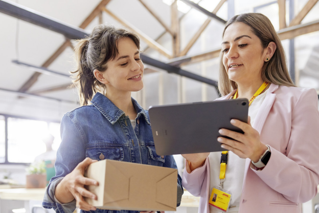 Two women are standing indoors. One holds a box while the other shows her something on a tablet. They are both smiling, likely discussing affordable small business shipping solutions perfect for their Small and Medium Business needs.