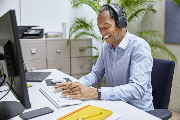 Una persona con auriculares y camisa azul sonríe mientras teclea en un escritorio con un monitor de ordenador, un cuaderno amarillo e información sobre envíos asequibles para pequeñas empresas.