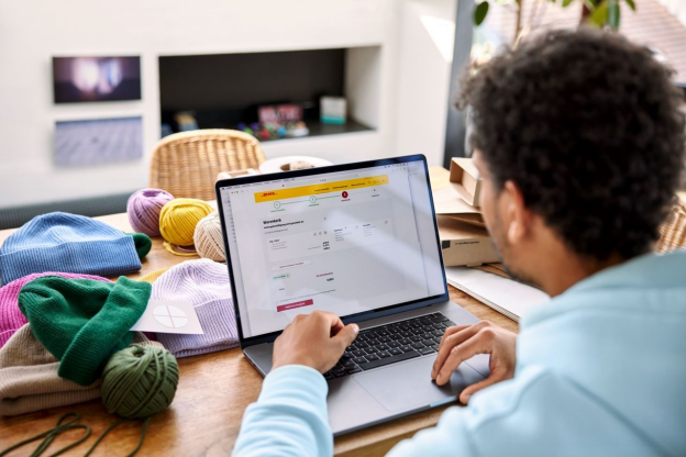 A person in glasses is using a laptop on a wooden table. Various balls of yarn and completed knitted hats are scattered around the table, highlighting their focus on creating handmade products while exploring small business shipping solutions.