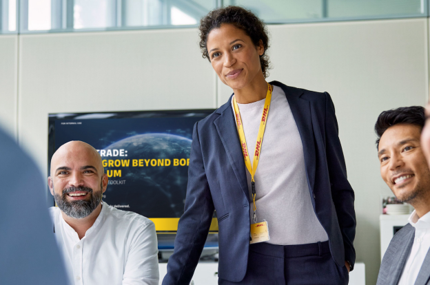 A woman in a business suit with a DHL lanyard stands and smiles in a meeting room. Three colleagues sit around her, all smiling. A screen in the background displays a presentation on affordable small business shipping solutions.