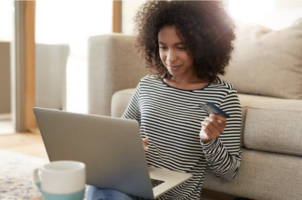 A woman with curly hair, wearing a striped shirt, sits on the floor using a laptop and holding a credit card, exploring shipping for small businesses.