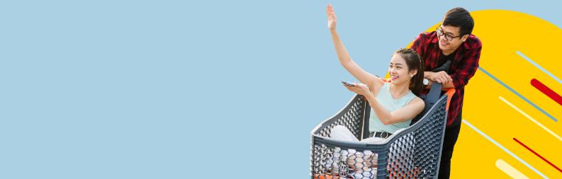 Happy man carrying an excited woman pointing upwards seated in a shopping cart.