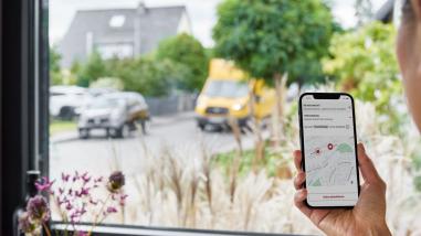 Woman looking out of a window holding a phone with the parcel tacking map on the phone. DHL eCommerce van outside driving towards the house.