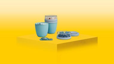 b.	Mepal food storage boxes and cups with food inside on a yellow plinth with a yellow background.