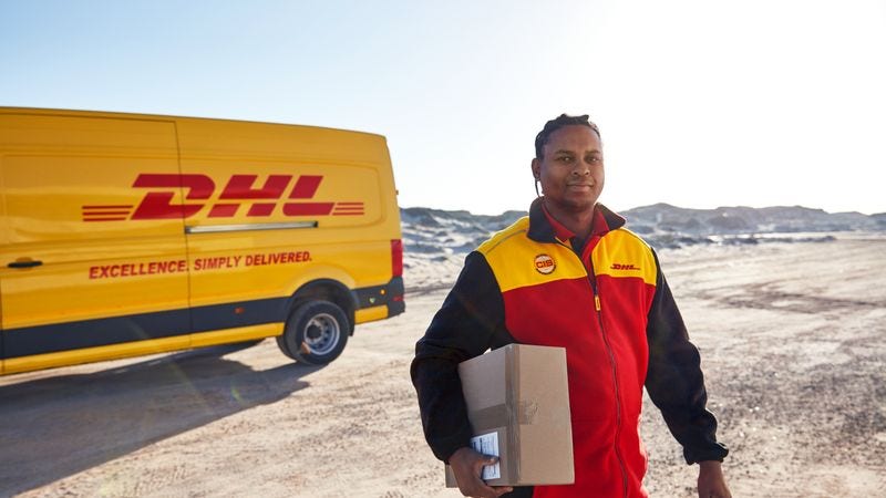 A DHL employee carries a parcel while walking on sandy ground with a delivery van in the background.