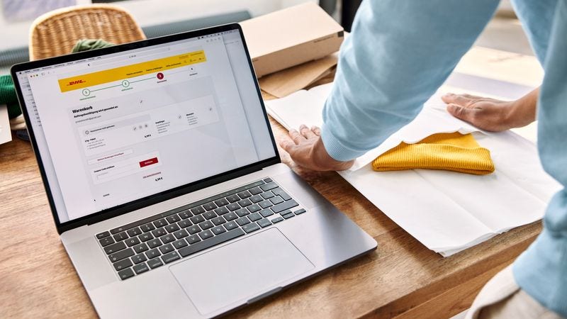 person preparing to ship a beanie on a table beside a laptop displaying a dhl shipping service webpage.