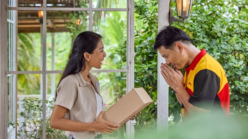A DHL Express courier greeting a customer while delivering a package at her doorstep in a lush, tropical setting.