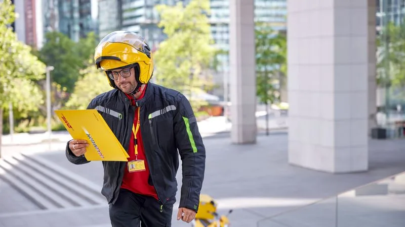 a dhl employee delivering an express envelope with his motorbike parked in the background