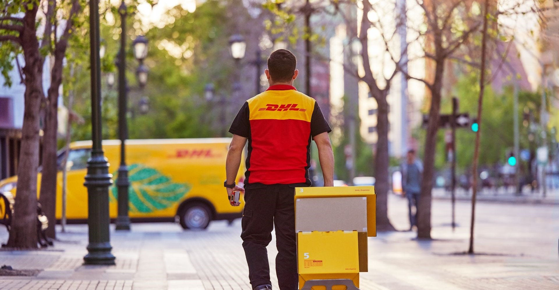 A DHL employee pulls a hand truck while walking towards a DHL GoGreen Plus van in a city.