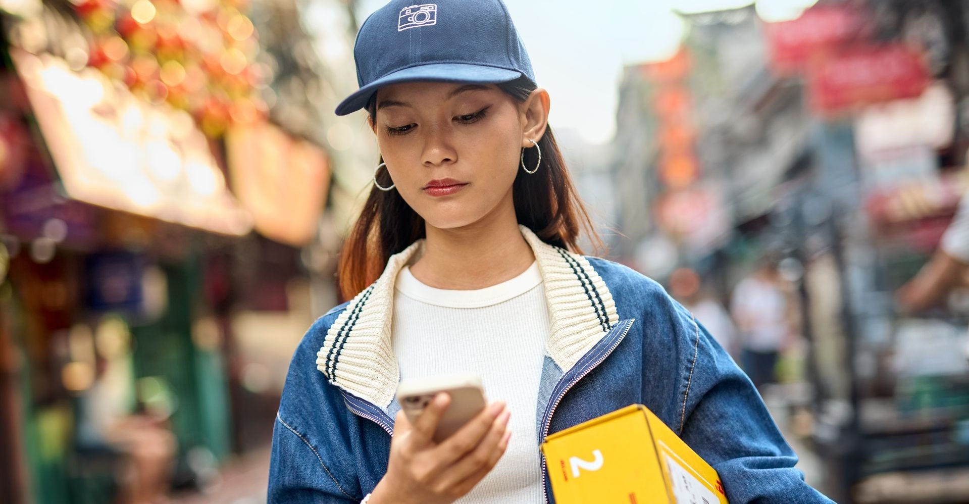 A young woman walks a busy street carrying a DHL express parcel under her arm and looking at her smartphone.