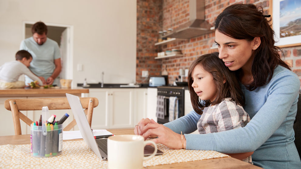 Woman sitting with her child on the laptop whilst husband makes food with son in the background