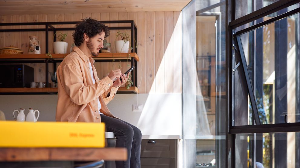 a male sitting on a kitchen counter, holding a smartphone with a dhl express parcel visible in the image