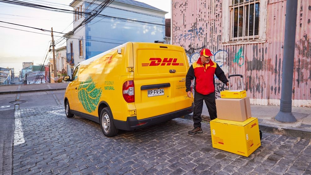 A male DHL employee is loading a hand truck with parcels next to his electric DHL van.